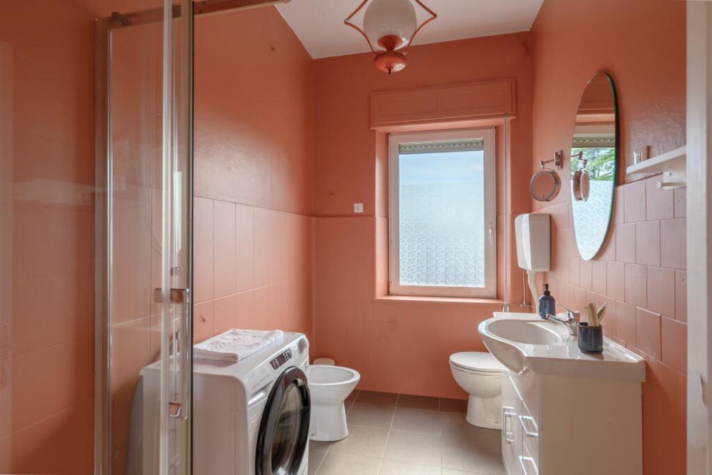 Vista su bagno dalla porta. Mattonelle color salmone, lavello, sanitari, lavatrice e doccia.