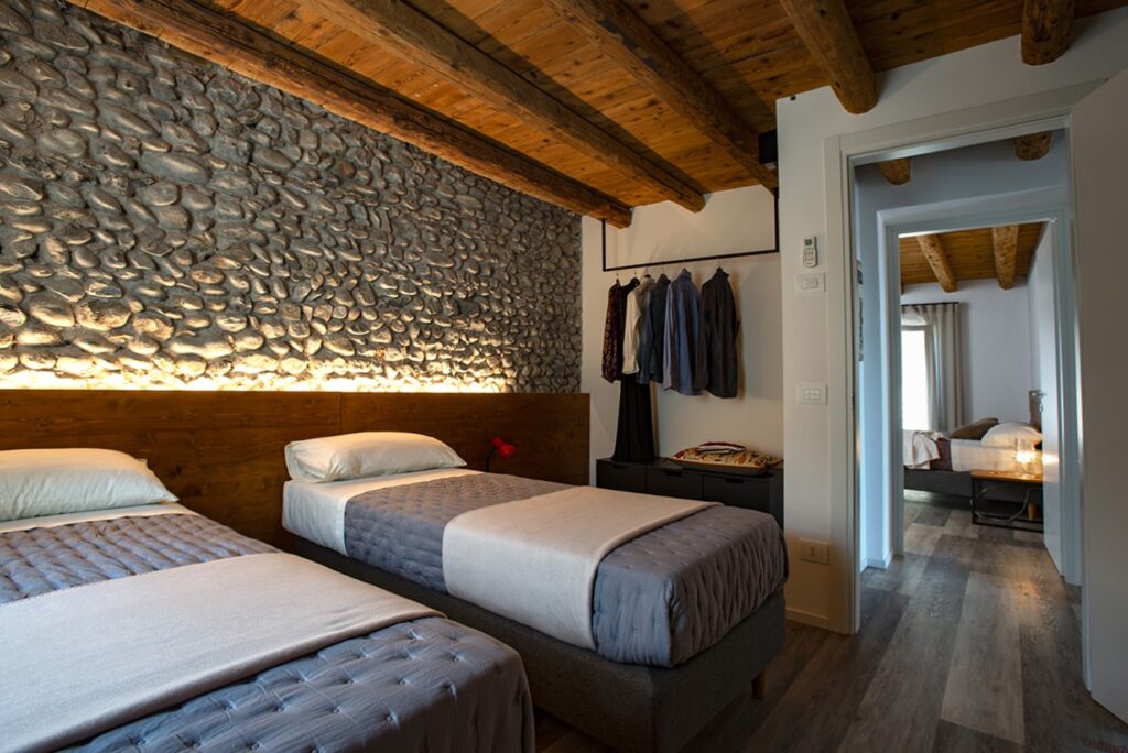 camera da letto moderna in ambiente rustico con due letti singoli, parete con pietre e soffitto a travatura